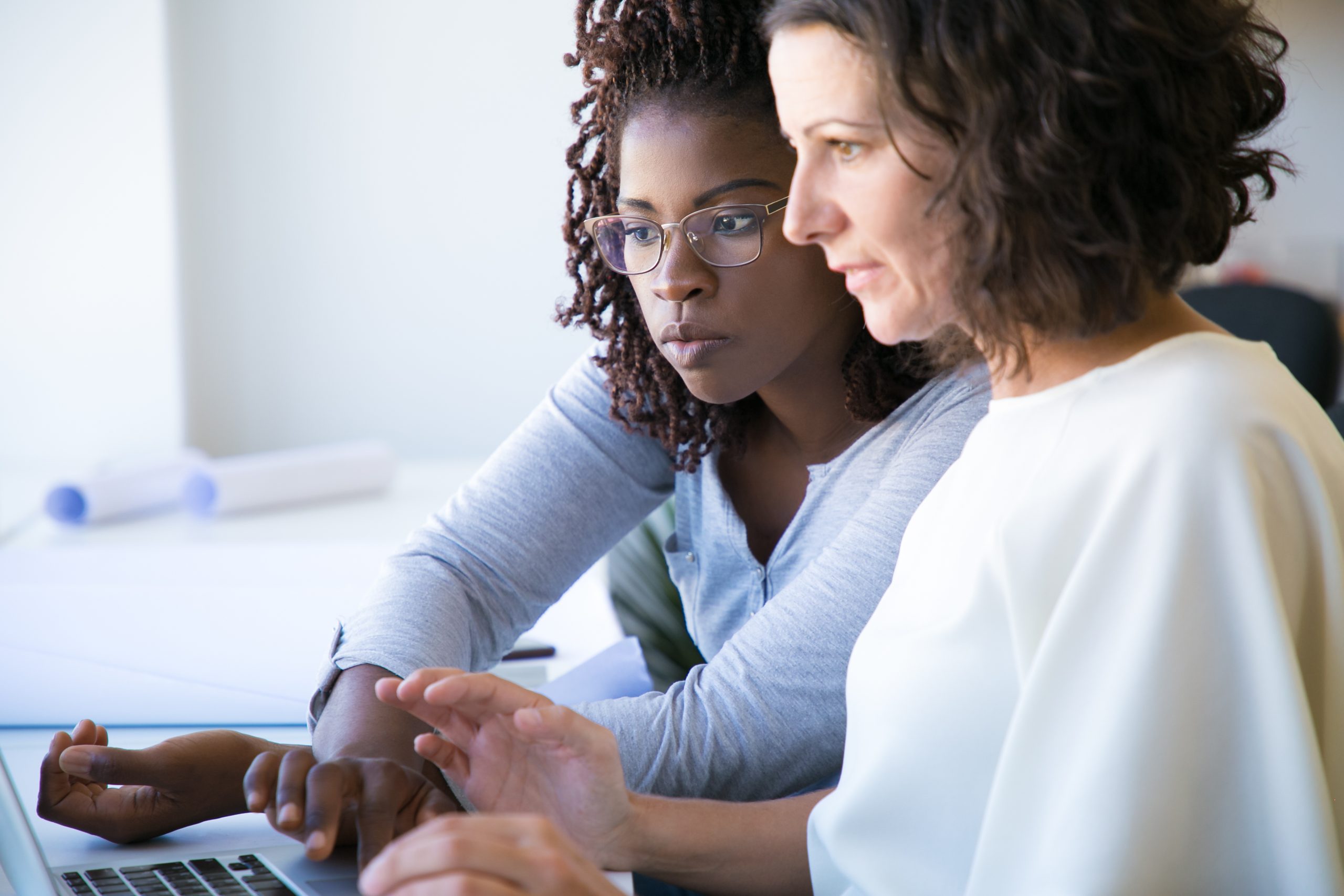 Female professional showing software specifics to colleague. Two diverse women using laptop in office. Corporate help concept